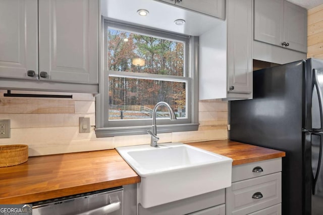 kitchen featuring tasteful backsplash, butcher block countertops, stainless steel dishwasher, sink, and black fridge