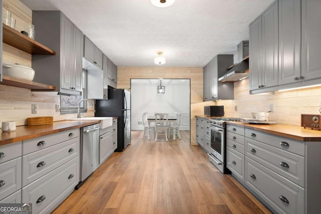 kitchen featuring appliances with stainless steel finishes, butcher block countertops, wall chimney exhaust hood, backsplash, and light wood-type flooring