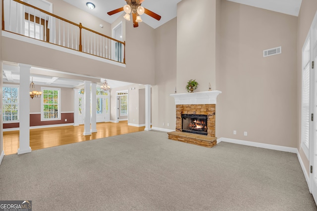 unfurnished living room featuring a high ceiling, ceiling fan with notable chandelier, a stone fireplace, carpet floors, and decorative columns