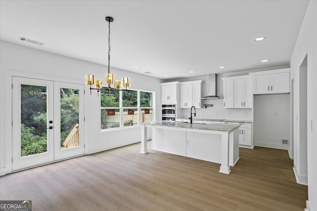 kitchen with sink, white cabinetry, light stone countertops, a kitchen island with sink, and wall chimney range hood