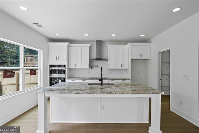 kitchen with wall chimney range hood, white cabinets, light stone counters, and stainless steel double oven