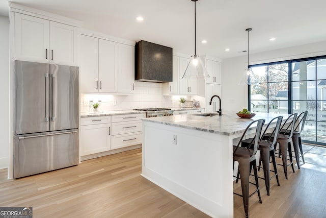 kitchen featuring sink, wall chimney exhaust hood, an island with sink, white cabinetry, and stainless steel refrigerator