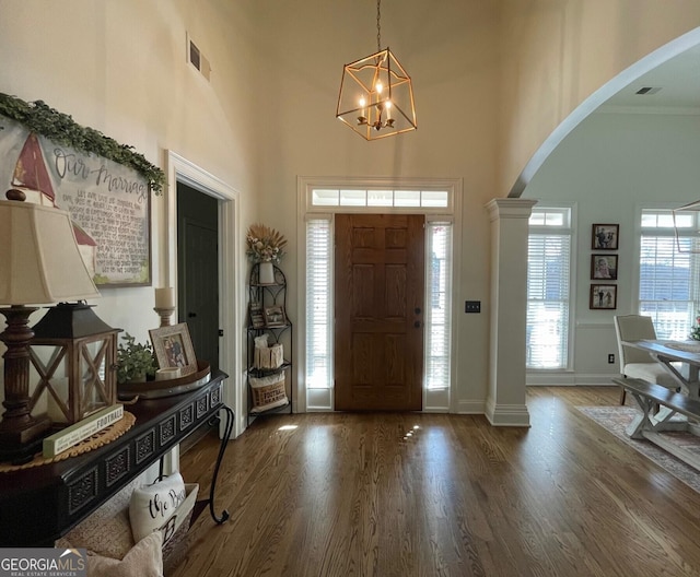 entryway featuring ornate columns, a towering ceiling, ornamental molding, a notable chandelier, and dark wood-type flooring