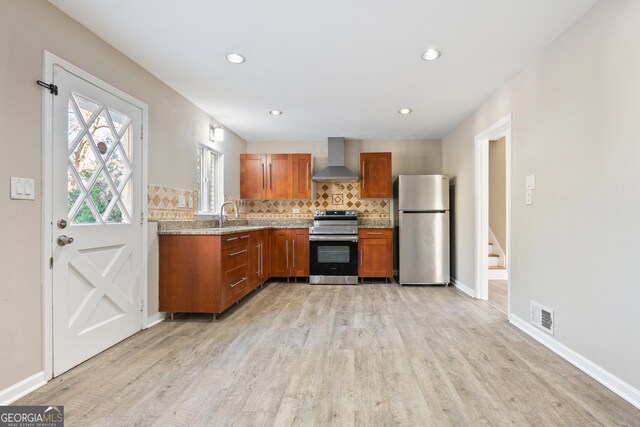 kitchen with wall chimney range hood, backsplash, appliances with stainless steel finishes, and light hardwood / wood-style flooring