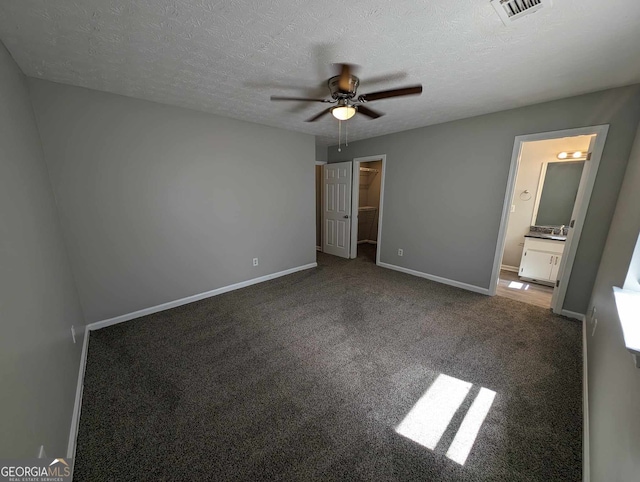 unfurnished bedroom featuring dark carpet, ensuite bathroom, ceiling fan, a spacious closet, and a textured ceiling