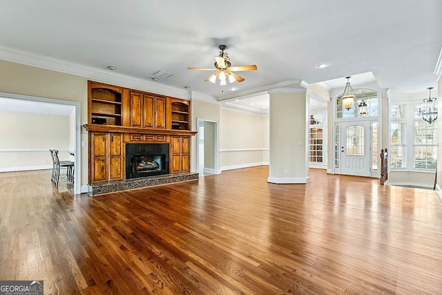 unfurnished living room featuring ceiling fan with notable chandelier, built in shelves, ornamental molding, a fireplace, and wood-type flooring