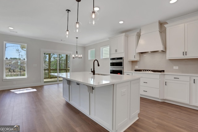 kitchen featuring appliances with stainless steel finishes, a center island with sink, a sink, light countertops, and custom range hood