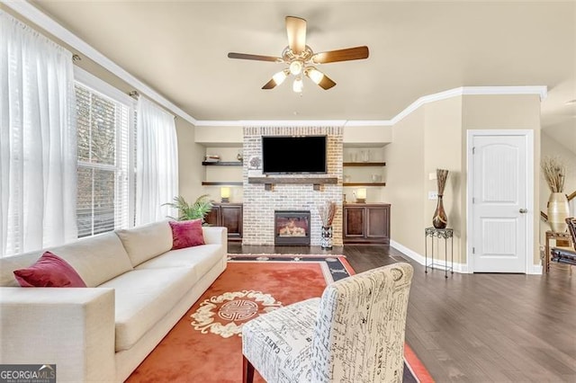 living area featuring ceiling fan, wood finished floors, baseboards, a brick fireplace, and crown molding