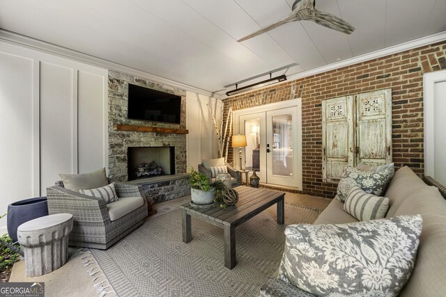 living room with ceiling fan, french doors, a stone fireplace, brick wall, and ornamental molding