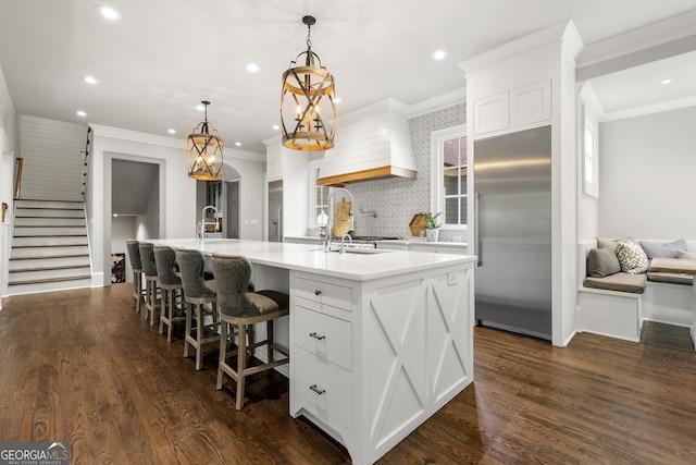 kitchen featuring dark wood-type flooring, hanging light fixtures, built in refrigerator, a kitchen island with sink, and white cabinets