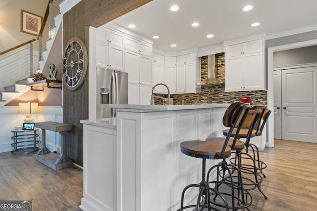 kitchen with ornamental molding, wall chimney range hood, light hardwood / wood-style flooring, white cabinets, and stainless steel fridge with ice dispenser