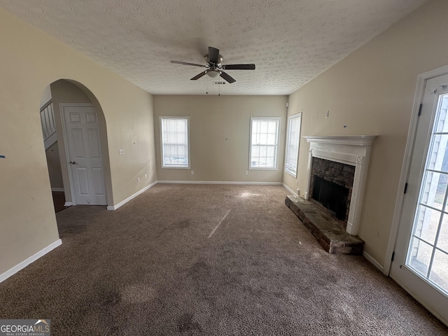 unfurnished living room featuring a textured ceiling, carpet floors, a stone fireplace, and ceiling fan