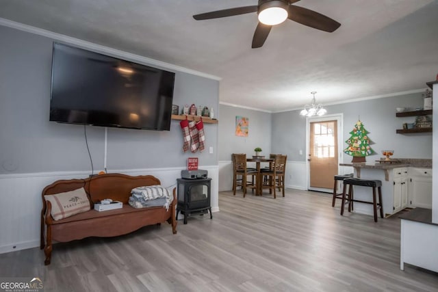 living room with ceiling fan with notable chandelier, light wood-type flooring, a wood stove, and crown molding