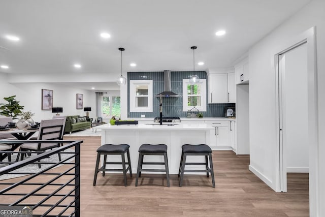 kitchen featuring a center island with sink, wall chimney exhaust hood, light wood-type flooring, tasteful backsplash, and white cabinetry