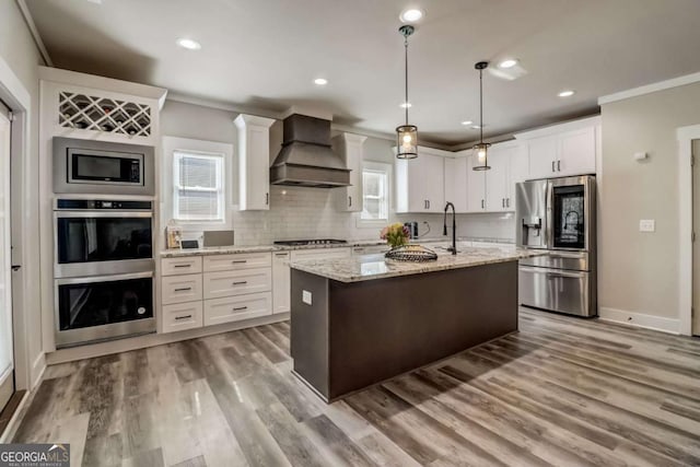 kitchen featuring wall chimney exhaust hood, light stone countertops, an island with sink, appliances with stainless steel finishes, and white cabinetry