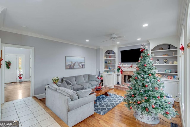 living room featuring light wood-type flooring, a brick fireplace, ornamental molding, built in shelves, and ceiling fan