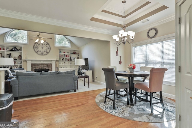 dining space featuring a raised ceiling, crown molding, light hardwood / wood-style floors, a fireplace, and ceiling fan with notable chandelier