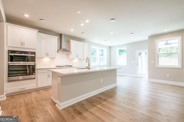 kitchen with double oven, white cabinets, a kitchen island with sink, and wall chimney exhaust hood