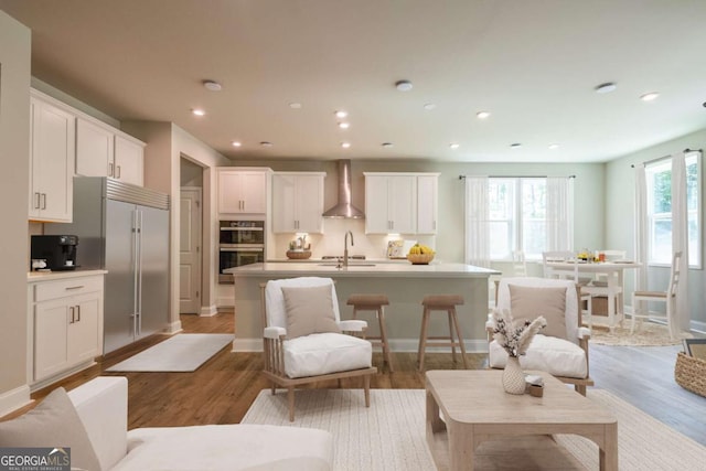 kitchen featuring wood-type flooring, a center island with sink, white cabinetry, and wall chimney range hood