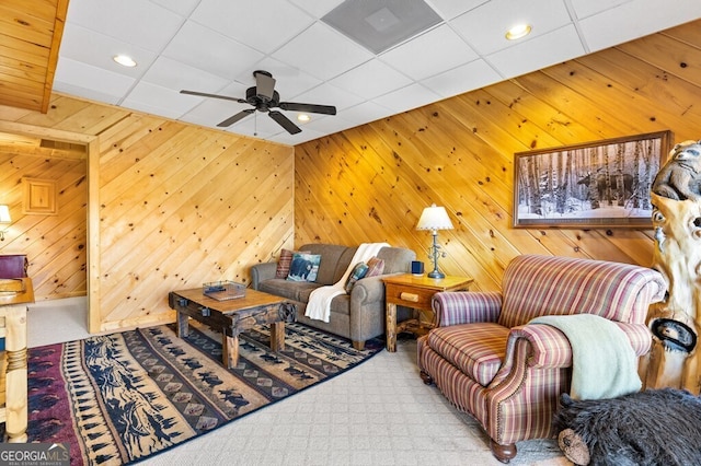 carpeted living room with a drop ceiling, wood walls, and ceiling fan