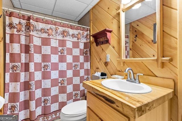 bathroom with a paneled ceiling, wooden walls, vanity, and toilet