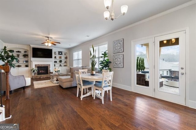 dining space with crown molding, ceiling fan with notable chandelier, dark hardwood / wood-style floors, built in shelves, and a stone fireplace