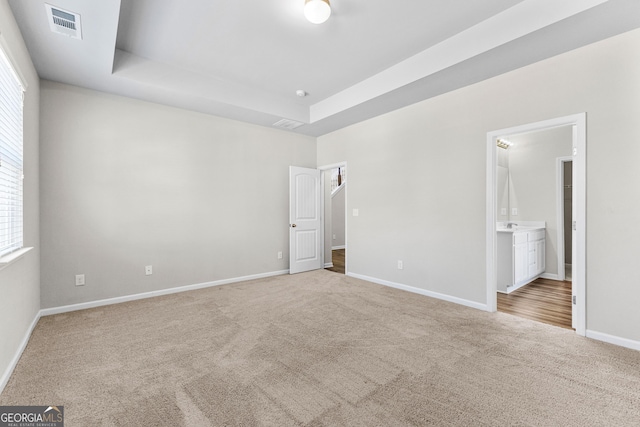 unfurnished bedroom featuring ensuite bathroom, light colored carpet, a tray ceiling, and multiple windows