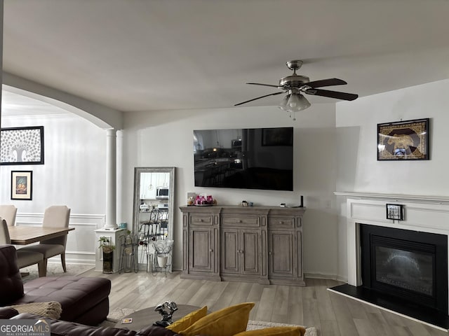 living area with arched walkways, decorative columns, a glass covered fireplace, ceiling fan, and light wood-type flooring