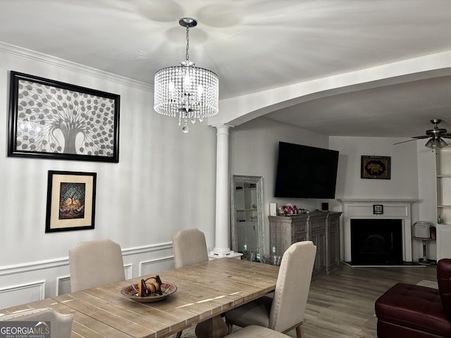 dining area featuring arched walkways, ceiling fan with notable chandelier, a fireplace, wood finished floors, and decorative columns