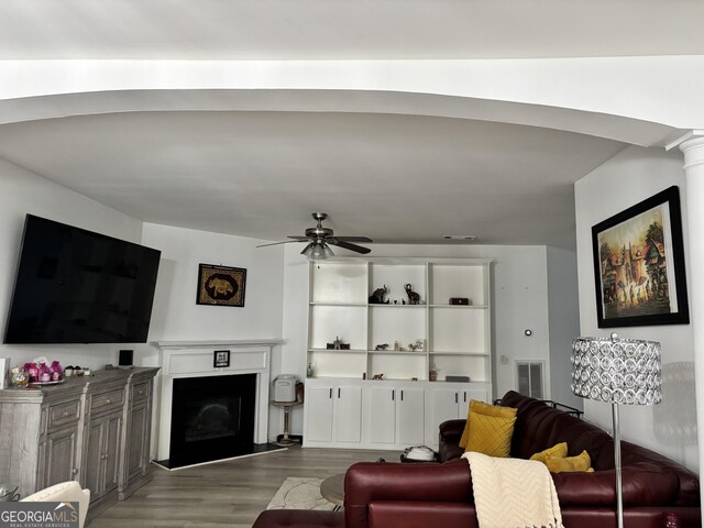 living room with visible vents, a fireplace with raised hearth, a ceiling fan, wood finished floors, and ornate columns