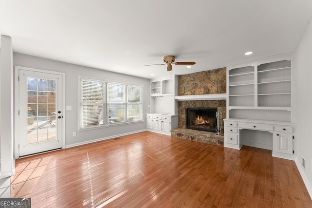 unfurnished living room with ceiling fan, built in shelves, a stone fireplace, and light hardwood / wood-style floors