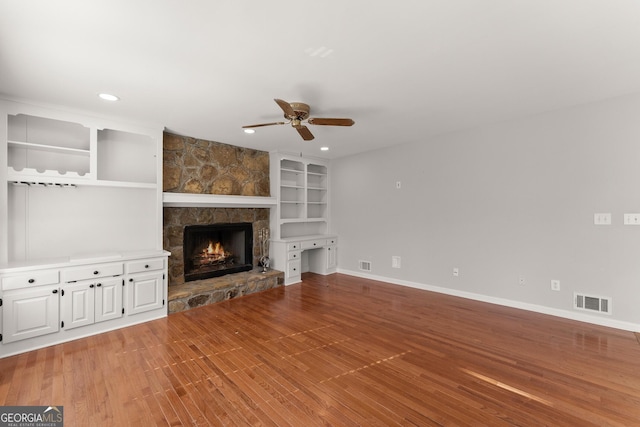 unfurnished living room with ceiling fan, built in features, a stone fireplace, and light hardwood / wood-style floors