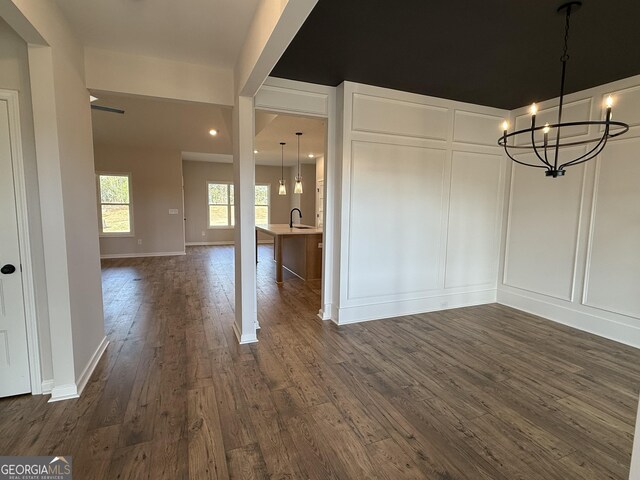 unfurnished dining area with dark wood-style flooring, a decorative wall, baseboards, a sink, and a notable chandelier