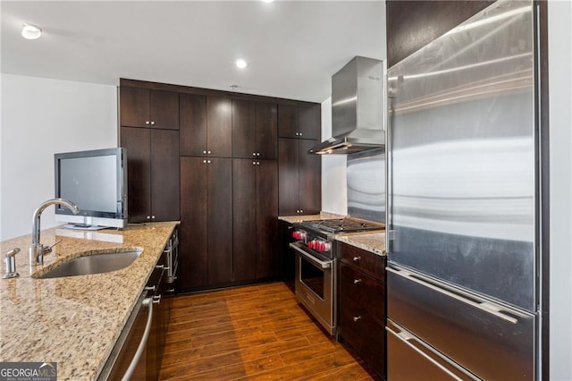 kitchen with light stone countertops, dark brown cabinetry, premium appliances, sink, and wall chimney range hood