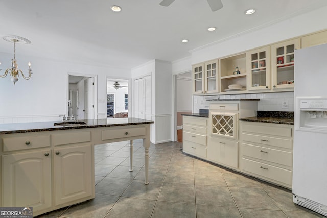 kitchen with white refrigerator with ice dispenser, pendant lighting, cream cabinetry, ceiling fan with notable chandelier, and tasteful backsplash