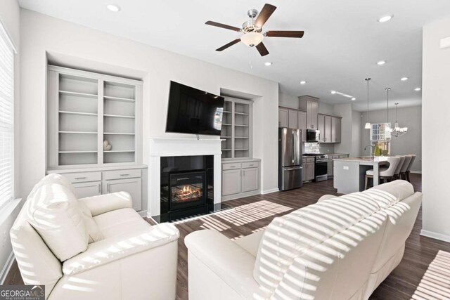 living room with built in shelves, dark wood-type flooring, and ceiling fan with notable chandelier