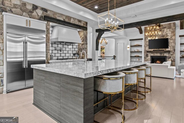 kitchen with custom exhaust hood, a stone fireplace, built in refrigerator, light stone countertops, and decorative light fixtures