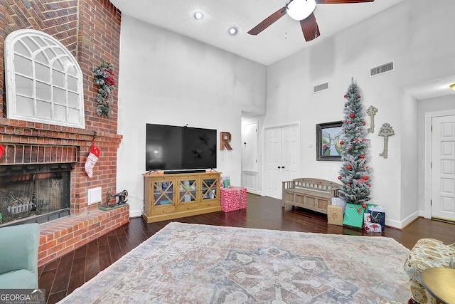 living room with ceiling fan, a fireplace, a towering ceiling, and dark wood-type flooring