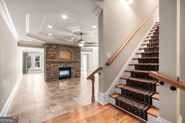 staircase featuring ornamental molding, a tray ceiling, ceiling fan, wood-type flooring, and a stone fireplace