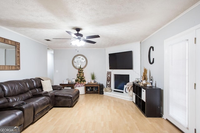 living room with a brick fireplace, a textured ceiling, ceiling fan, crown molding, and light hardwood / wood-style flooring