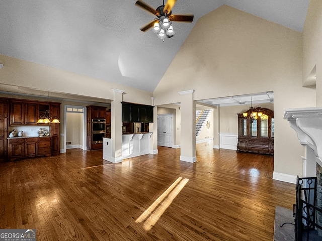 unfurnished living room with ceiling fan with notable chandelier, high vaulted ceiling, dark hardwood / wood-style floors, and ornate columns