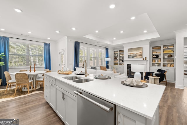 kitchen with stainless steel dishwasher, a tray ceiling, sink, hardwood / wood-style flooring, and a center island with sink