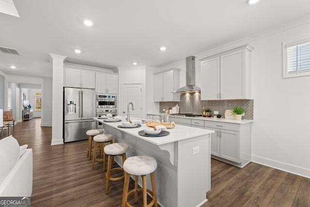 kitchen featuring white cabinetry, wall chimney exhaust hood, stainless steel appliances, a kitchen breakfast bar, and an island with sink