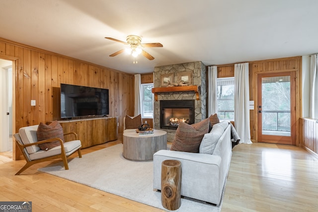 living room featuring ceiling fan, light hardwood / wood-style floors, a stone fireplace, and wooden walls