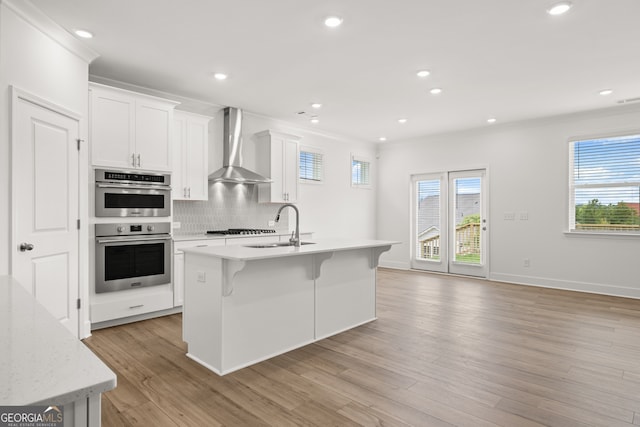 kitchen with white cabinetry, sink, wall chimney exhaust hood, stainless steel double oven, and a center island with sink