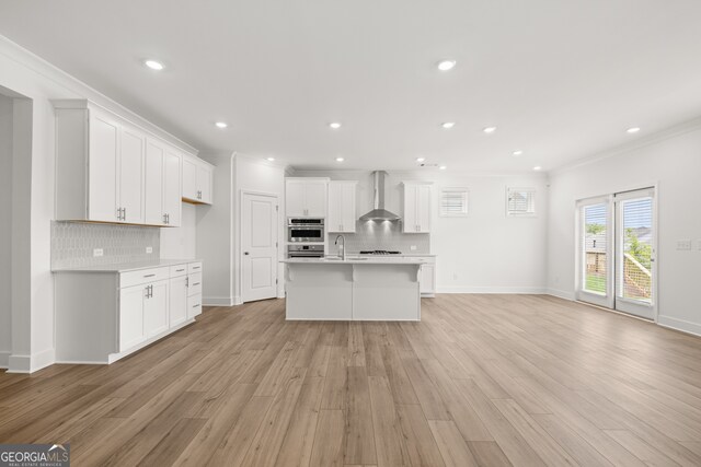 kitchen with a kitchen island with sink, wall chimney range hood, crown molding, tasteful backsplash, and white cabinetry