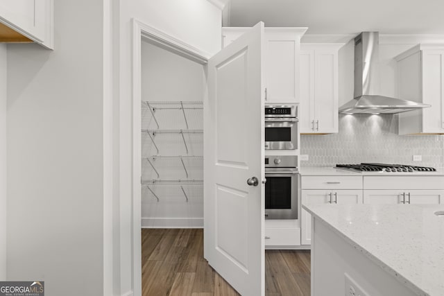 kitchen featuring wall chimney range hood, hardwood / wood-style flooring, light stone countertops, white cabinetry, and stainless steel appliances