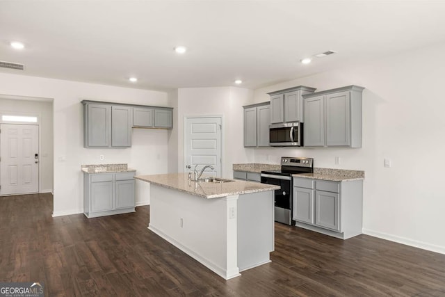 kitchen with light stone countertops, appliances with stainless steel finishes, dark hardwood / wood-style flooring, gray cabinetry, and a center island with sink