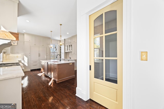 kitchen with light stone countertops, dark hardwood / wood-style flooring, pendant lighting, decorative backsplash, and a center island with sink