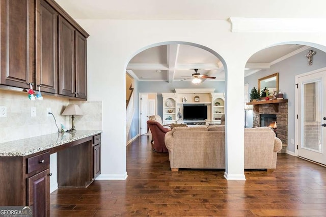 living room with beam ceiling, ceiling fan, dark wood-type flooring, coffered ceiling, and a stone fireplace